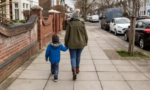 mother and child walking away on footpath - brick fences to the left, parked cars on the right