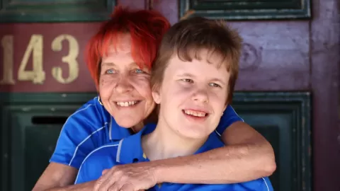 mother and son facing camera and smiling. Mother has arm around son (from behind)